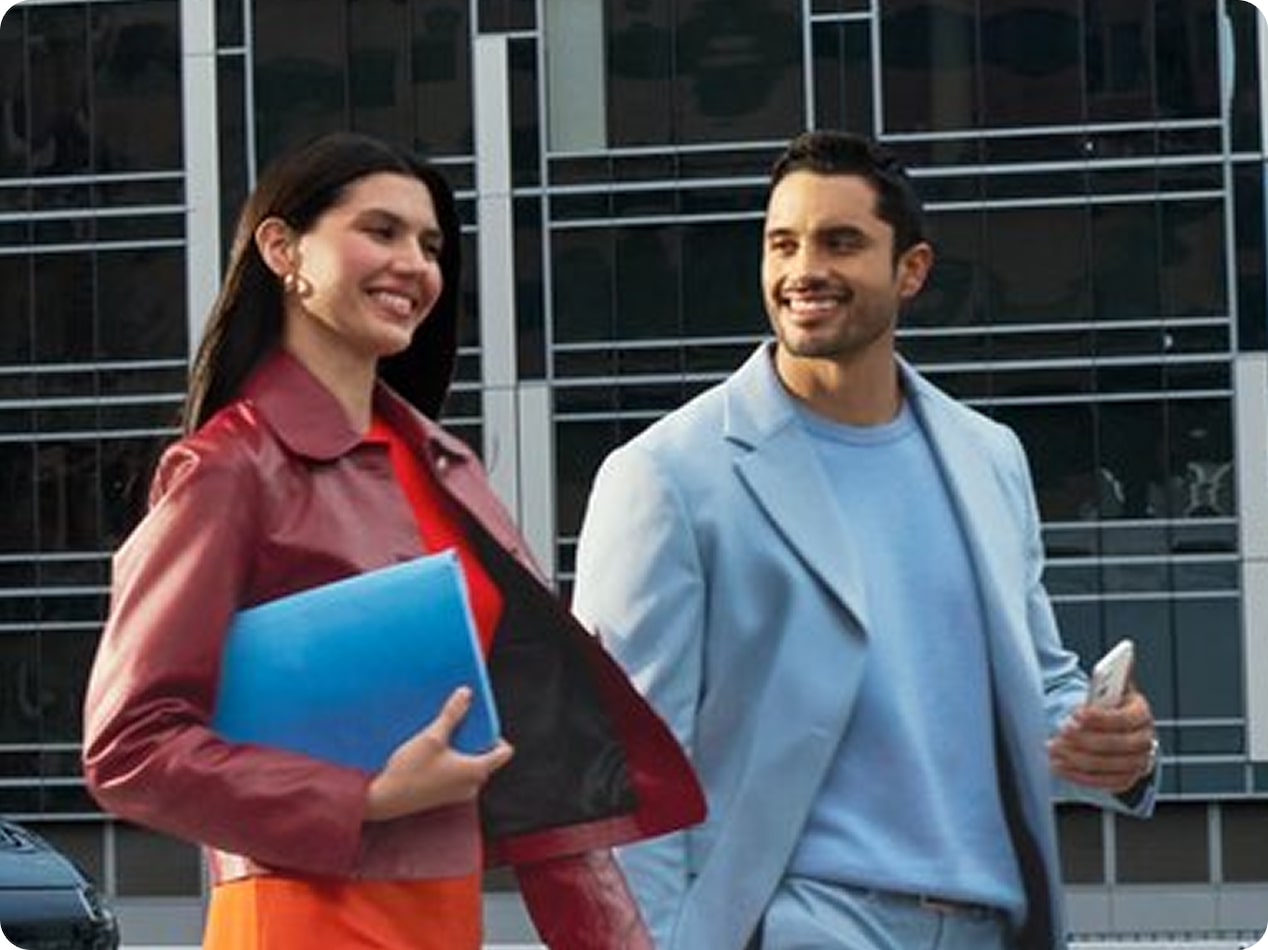 A man and woman dressed in business casual attire smile while walking past a modern glass building, representing the connected lifestyle and safety services provided by OnStar and HondaLink.
