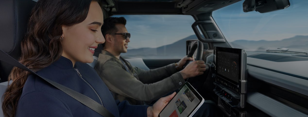 A woman in the passenger seat smiles while using a tablet as a man drives a vehicle through a mountainous landscape, highlighting the in-vehicle connectivity and convenience of OnStar.