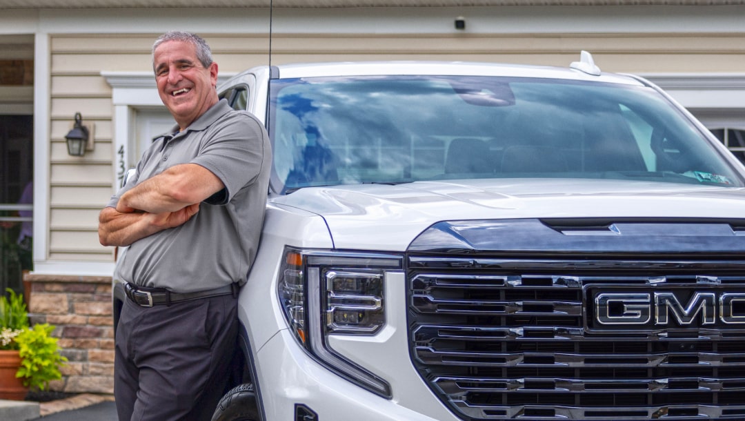 A man wearing a gray polo shirt and black pants leans against the grille of a white GMC Sierra pickup truck outside a beige house.