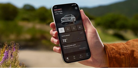 A person's hand holds a black smartphone displaying the GMC mobile app showing the vehicle's remote start and climate control features against a blurred background in an outdoor setting.