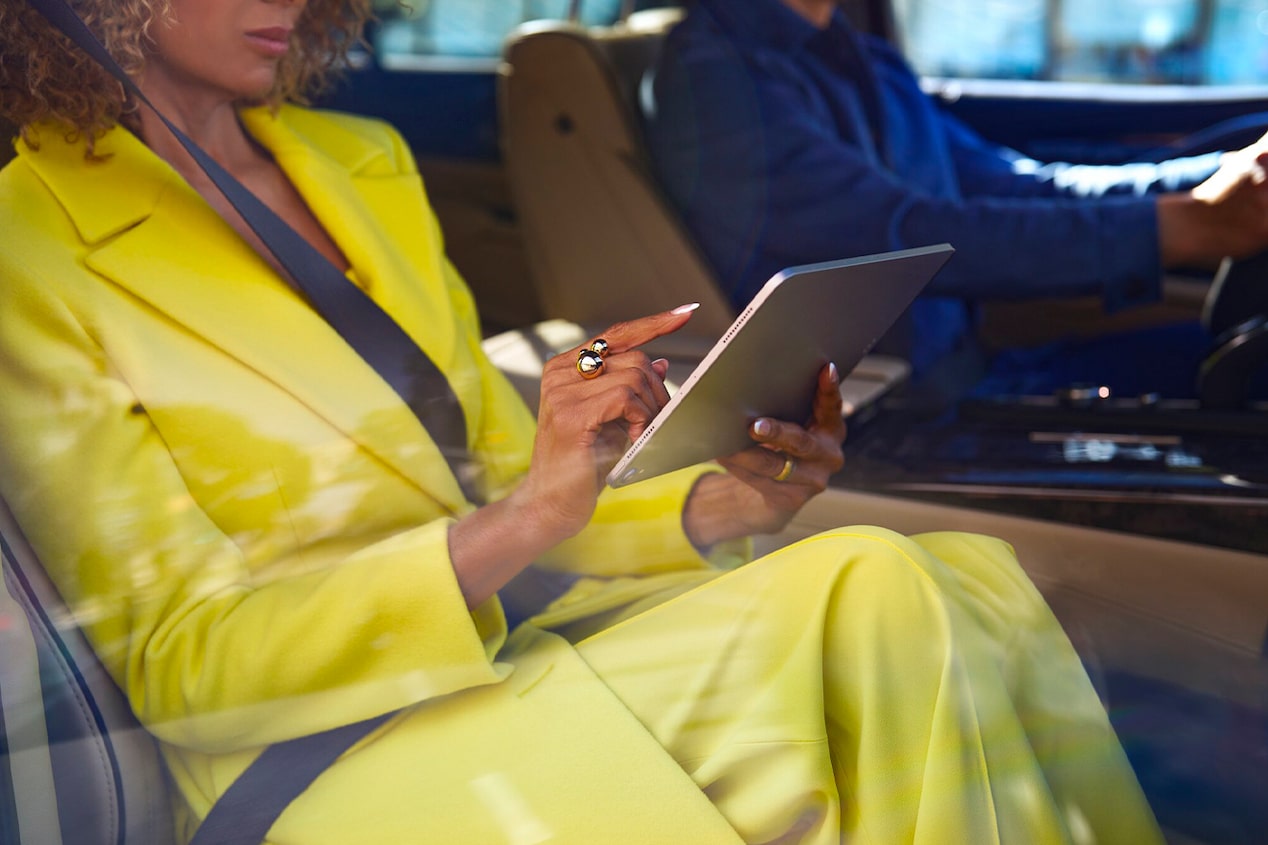 A close up view of a woman in a yellow pantsuit using a tablet and a man in a blue shirt in the driver's seat.