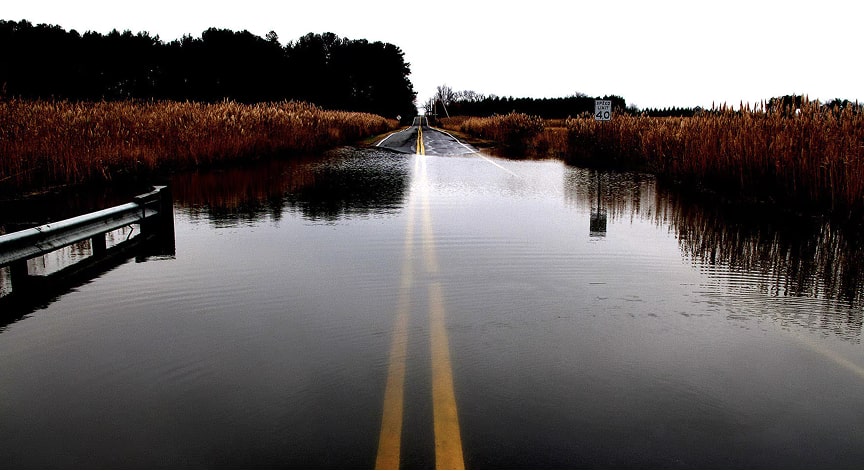 A straight paved road is heavily flooded with water, flanked by brown reeds under a cloudy sky.