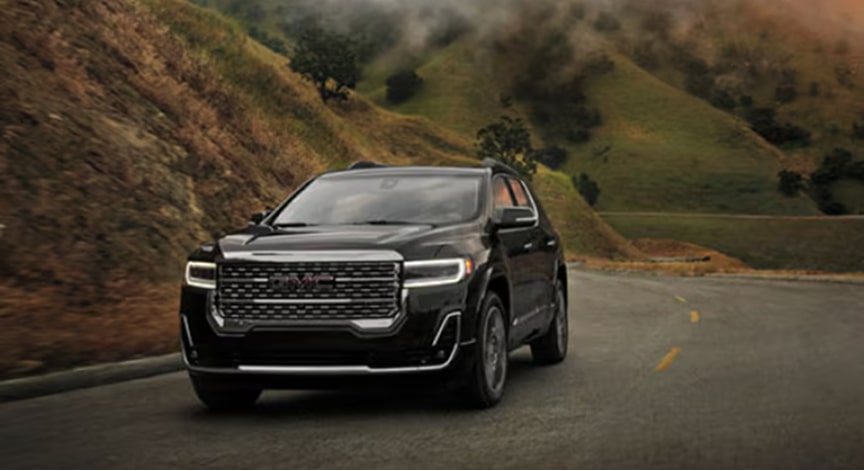 A black GMC Acadia SUV drives on a winding mountain road with foggy peaks in the background.