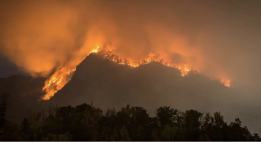 A large wildfire blazes across a dark mountainside, casting an orange glow into the night sky.