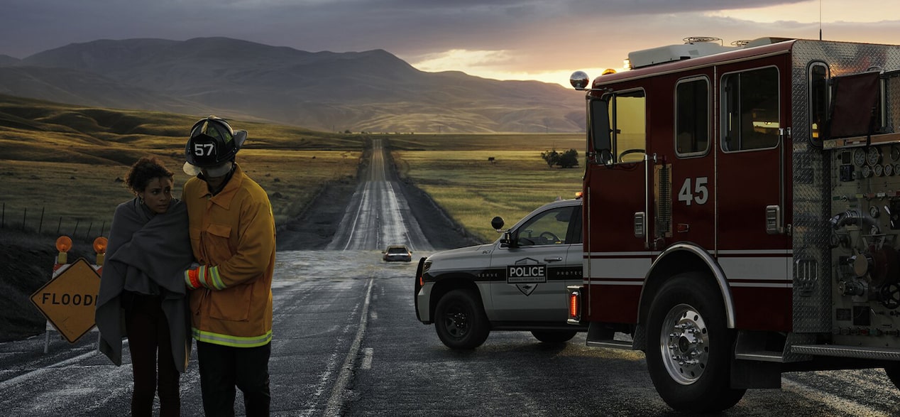 A firefighter helps a person wrapped in a blanket on a wet road next to a fire truck and police car with a flood warning sign.