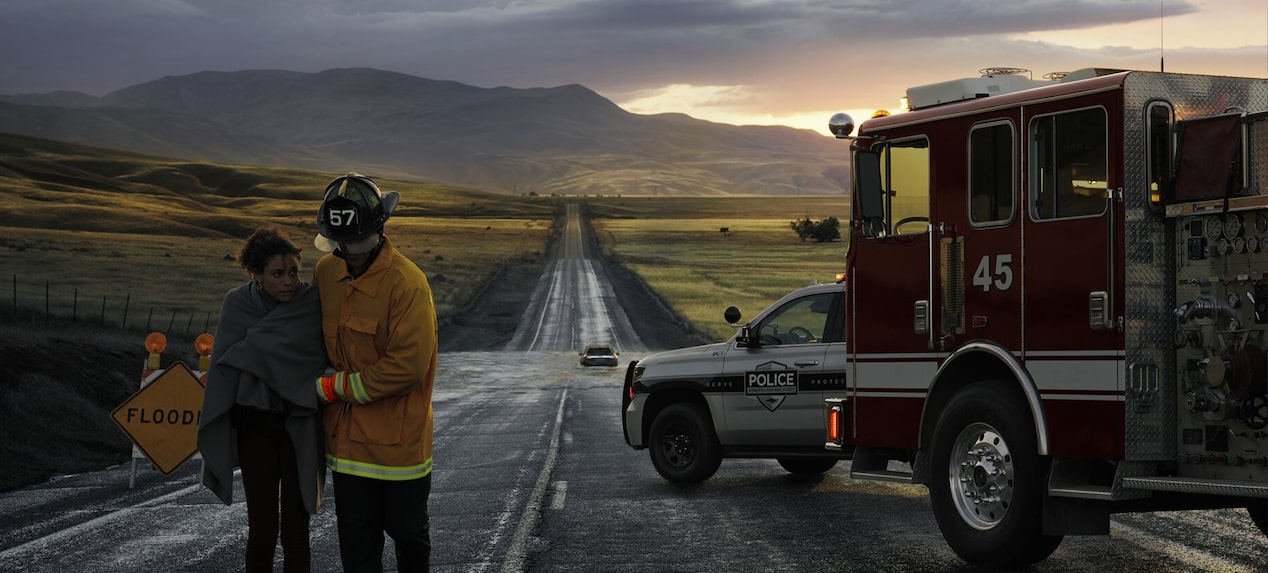 A firefighter helps a person wrapped in a blanket on a wet road next to a fire truck and police car with a flood warning sign.