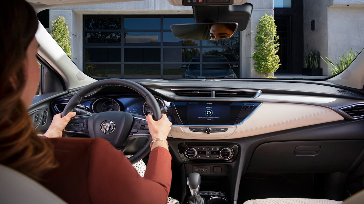 The interior view from the driver's seat of a Buick, showing a woman's hands on the steering wheel while the infotainment screen displays the Amazon Alexa "Speaking" interface, with a garage door visible through the windshield.