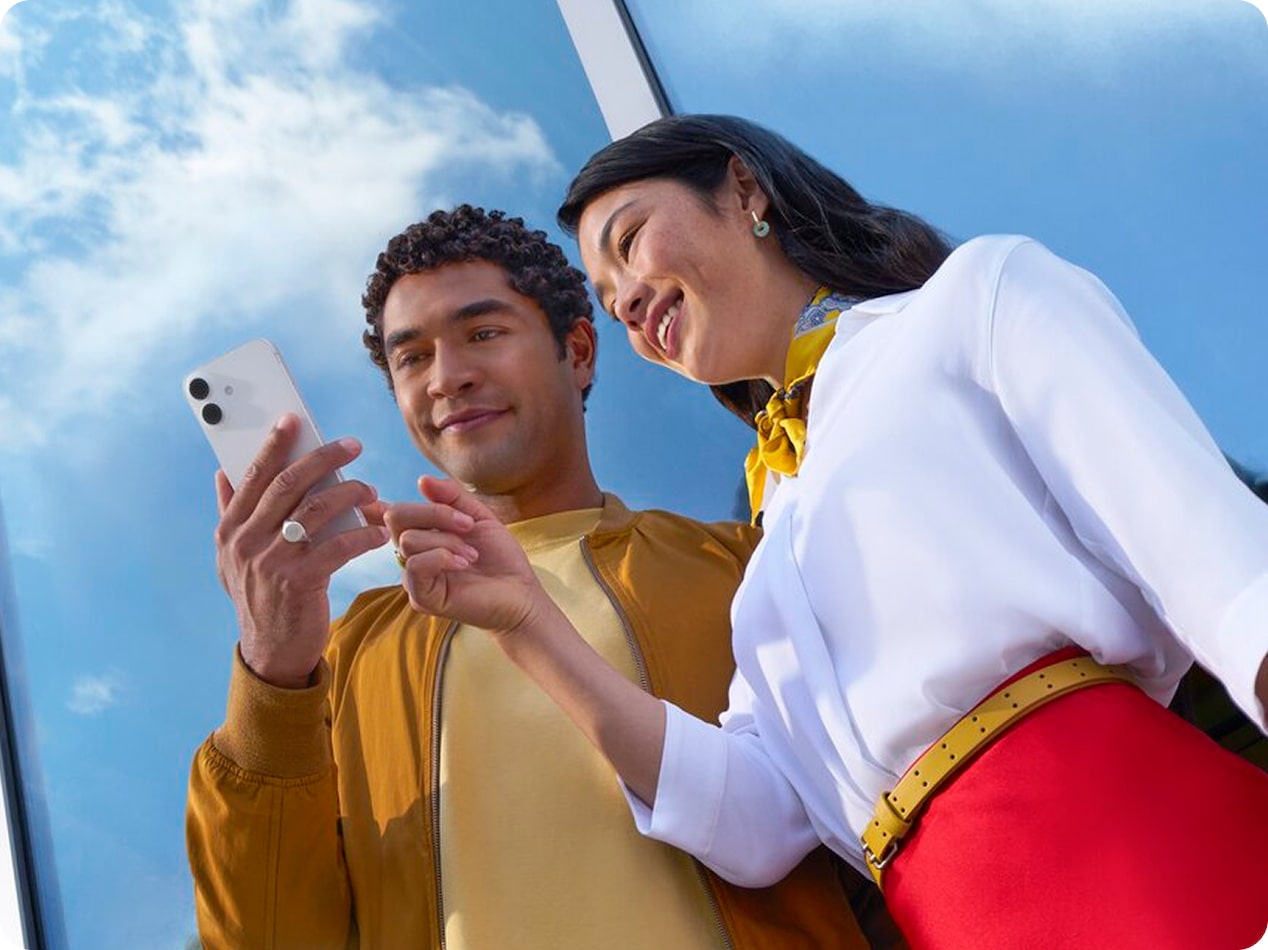 Two smiling people look at a smartphone, pointing at the screen under a bright blue sky.
