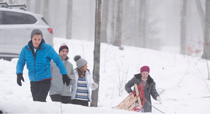 A group of people dressed in winter gear pulls a sled through deep snow next to a silver SUV in a snowy forest.