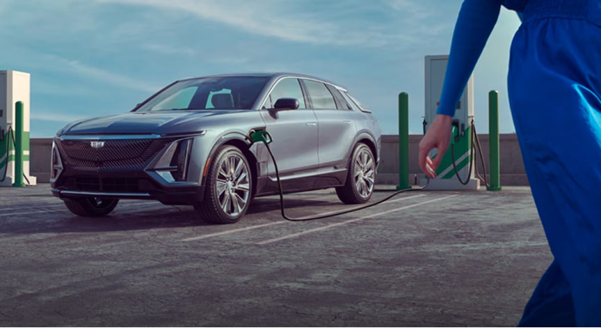 A silver Cadillac Lyriq electric vehicle charges at a public charging station as a person in blue walks past.