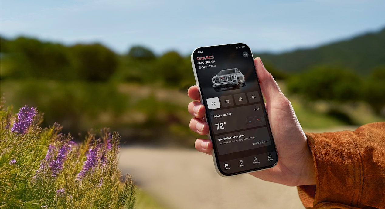 A person's hand holds a black smartphone displaying the GMC mobile app showing the vehicle's remote start and climate control features against a blurred background in an outdoor setting.