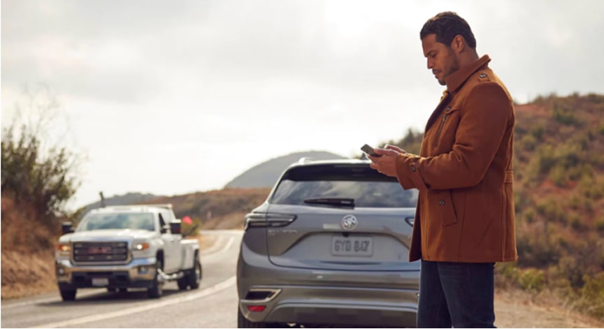 Man standing by a silver SUV and a GMC tow truck on the side of a rural road, using a phone.