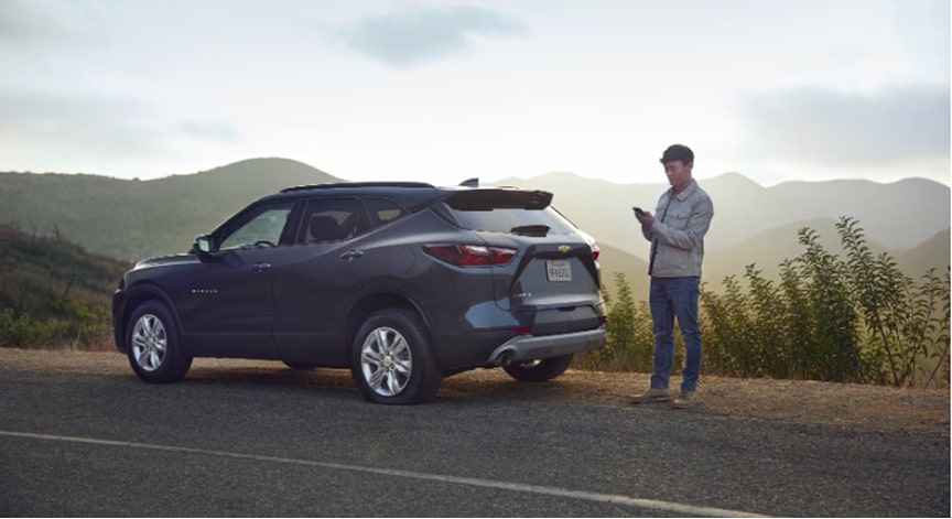 Man standing beside a black Chevrolet Blazer on the side of a rural road, using a phone to request OnStar roadside asistance.