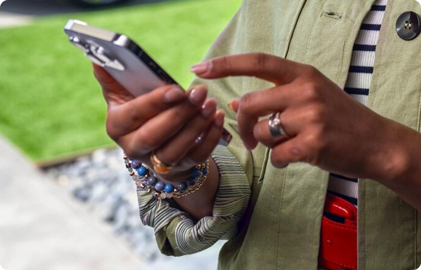 Close-up of a person's hands holding and interacting with a silver smartphone outdoors to request the stolen vehicle OnStar assistance, with green grass in the background.
