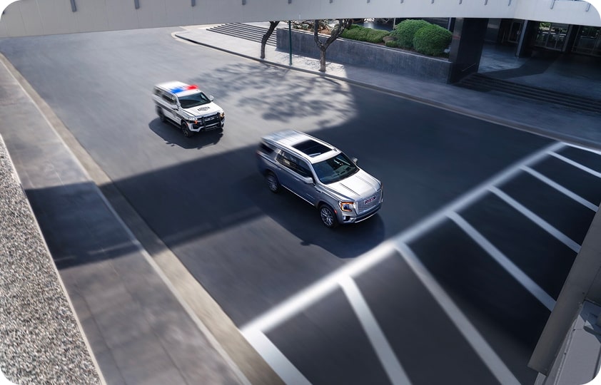 An overhead view of a silver SUV driving on a city street with a police car following behind.