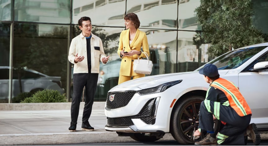 A person in a high-visibility vest changing a tire on a white Cadillac sedan, with two people watching.