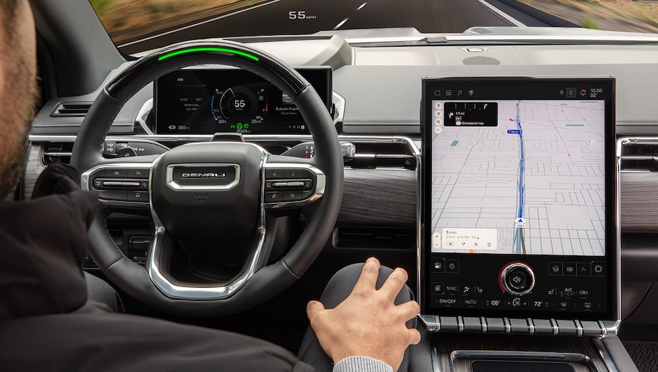 A man sits in a GMC car with hands on his legs, the steering wheel shows a green light indicating that Super Cruise Hands Free Driving is active, and a large central screen displays a navigation map.