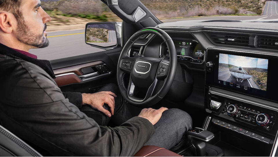 A bearded man sits in a GMC truck with a green light on the steering wheel indicating that Super Cruise Hands Free Driving is active, and the infotainment screen shows a rearview camera view of a trailer.