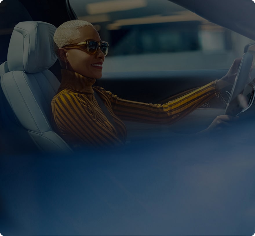 A smiling woman with short blonde hair and sunglasses is driving a car.