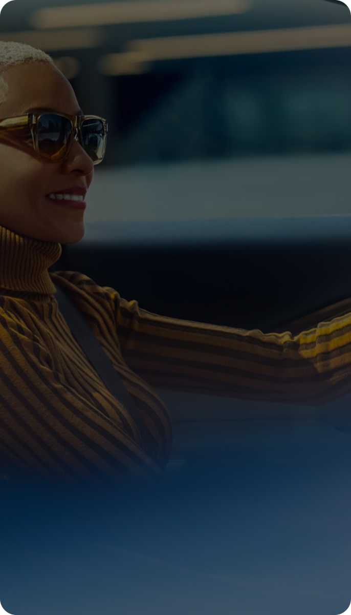 A smiling woman with short blonde hair and sunglasses is driving a car.