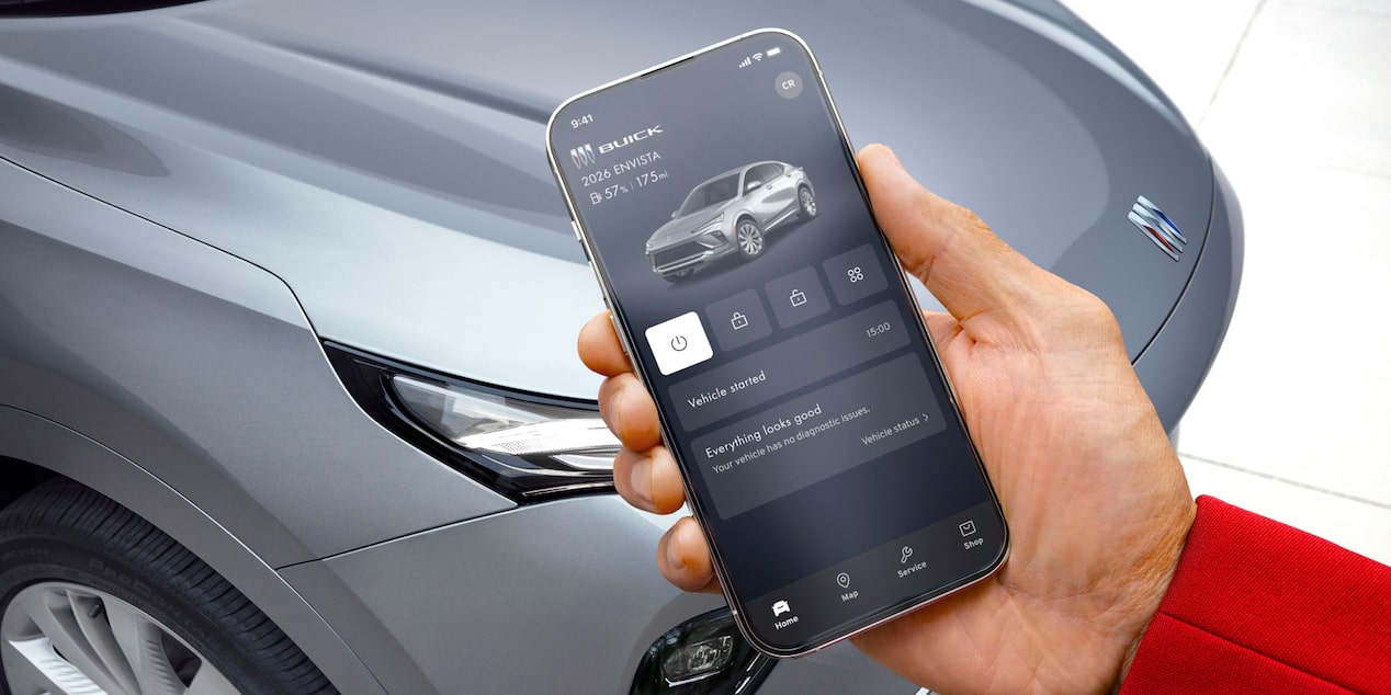  A hand holds a smartphone displaying the Buick mobile app with remote vehicle controls with a grey Envista 2026 in the background of an outdoor setting.