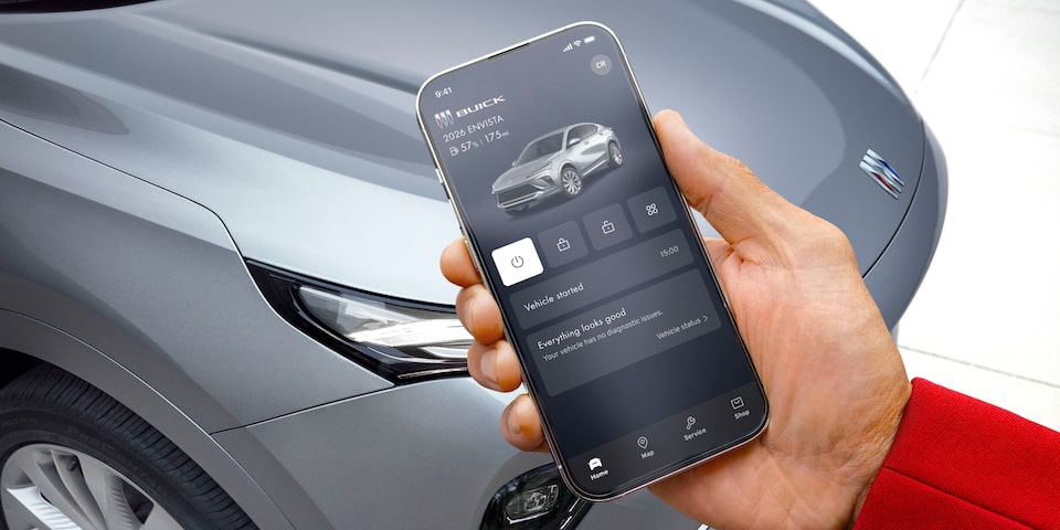  A hand holds a smartphone displaying the Buick mobile app with remote vehicle controls with a grey Envista 2026 in the background of an outdoor setting.