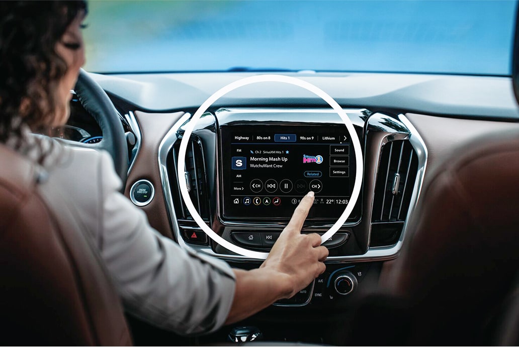 A person's hand touches the center display screen of a car, which shows the SiriusXM interface.