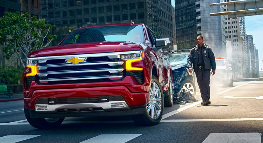 A red Chevrolet Silverado truck is parked on a city street with a police officer walking near a damaged car in the background.