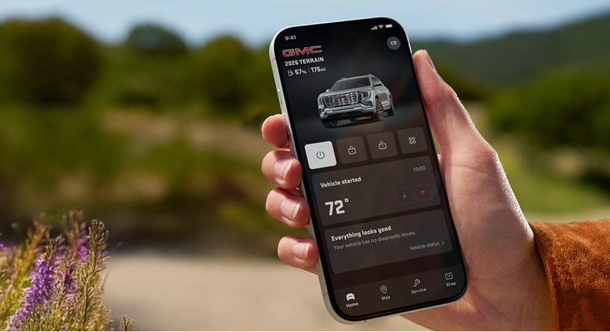 A person's hand holds a black smartphone displaying the GMC mobile app showing the vehicle's remote start and climate control features against a blurred background in an outdoor setting.