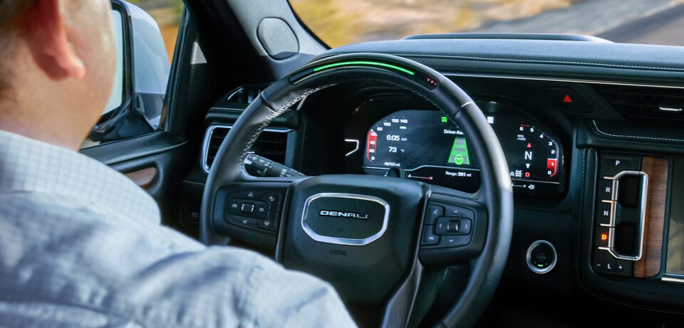 An over-the-shoulder shot of Milas R. driving a GMC Denali, with a green light on the steering wheel indicating that the hands-free Super Cruise feature is active.