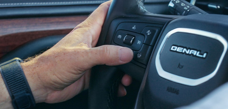 A close-up shot of Milas R.'s hand with a watch on his wrist pressing a button on the steering wheel of the GMC Denali, activating a function of the vehicle's driver assistance technology.