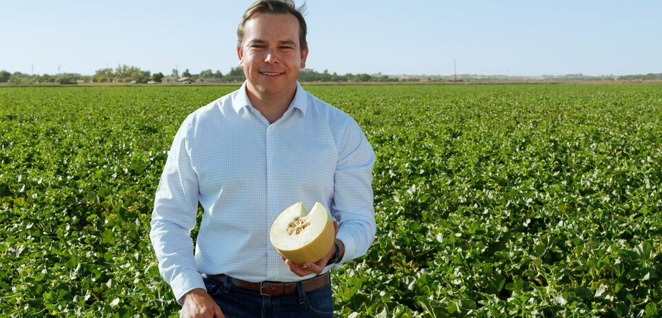 A smiling man, Milas R., stands in a field holding a large, freshly cut melon half in his hand.