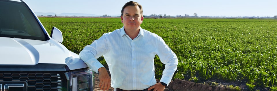 A smiling man, Milas R., in a white button-down shirt is standing in a field of green crops, leaning against the front of his white GMC Yukon with hands-free driver assistance technology from OnStar.