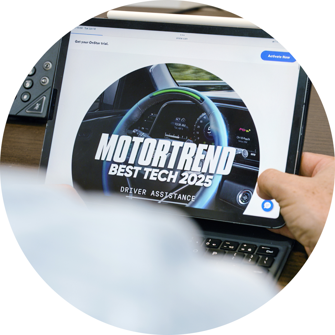 A hand holds a tablet displaying an article from Motortrend with the headline "Best Tech 2025" and a circular image of a GMC vehicle's steering wheel with the Super Cruise light bar glowing green.