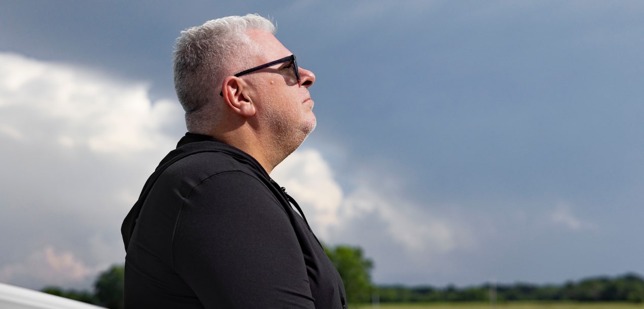 A profile view of Jason Cupp wearing sunglasses looking up at a dark, stormy sky over a green field.
