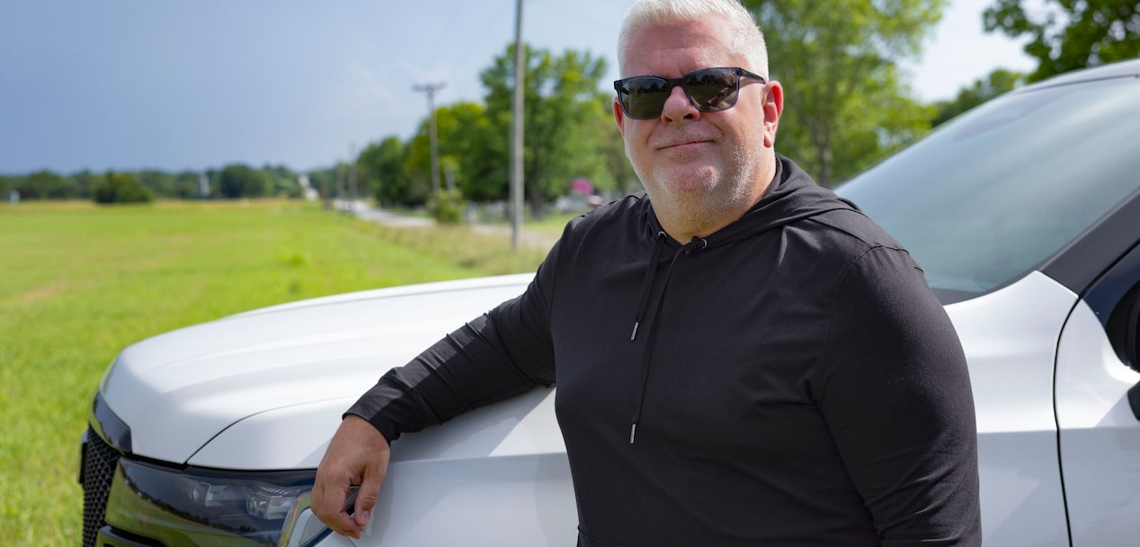 Jason Cupp leans casually against the hood of a silver suv on a rural road with a green field behind him.