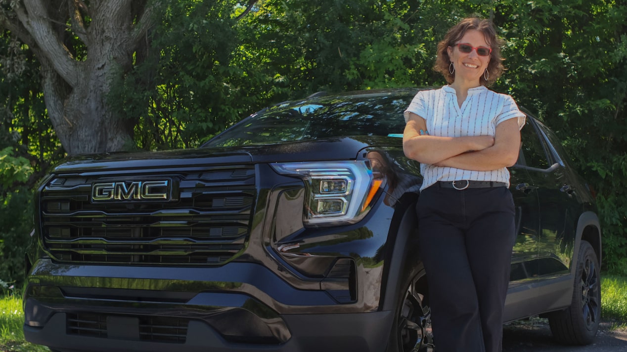 Kristy K, smiling with crossed arms, stands proudly in front of her black GMC Terrain, showcasing connected vehicle safety.