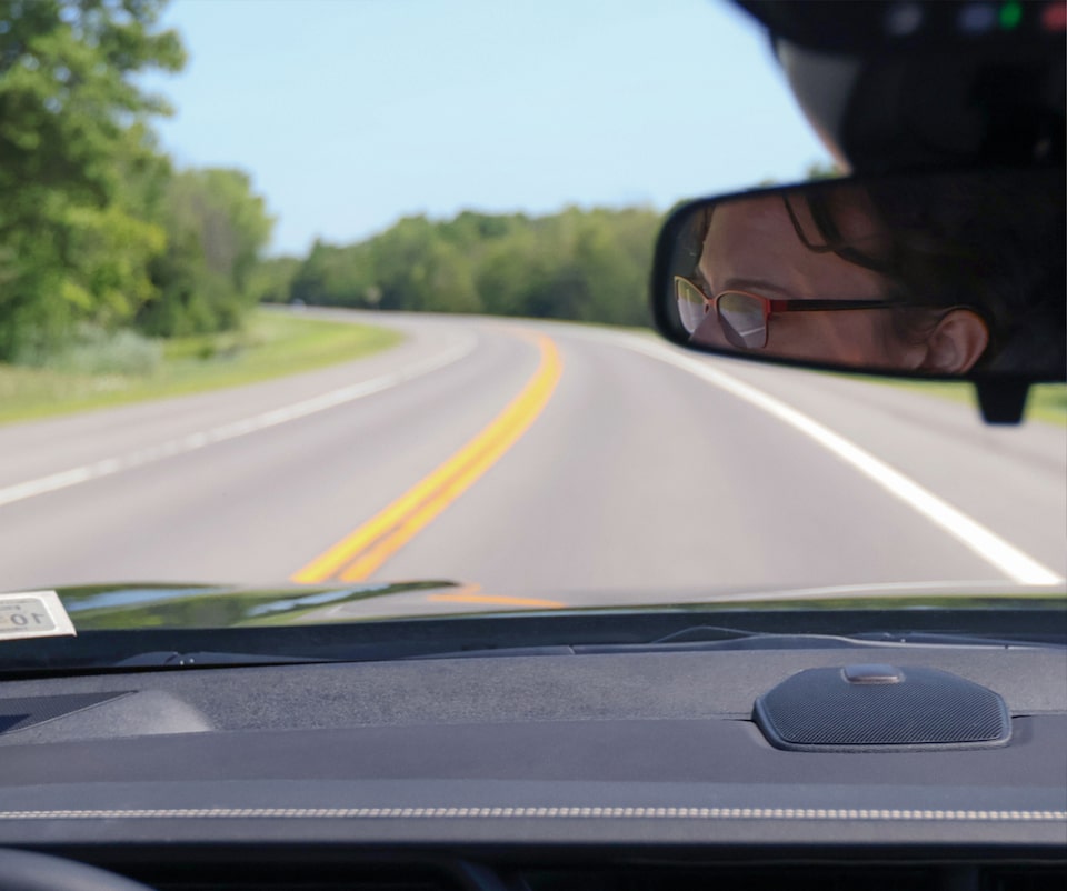A driver's perspective from inside a vehicle on a winding road, with Kristy K's reflection visible in the rearview mirror, representing driver safety services.