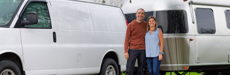 A man and a woman standing next to each other in front of their van and camper.