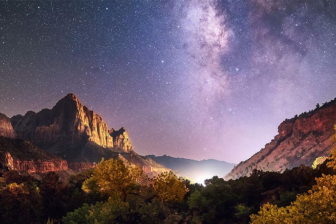 A night sky filled with stars and the Milky Way stretches above towering rock formations and a mountainous landscape.