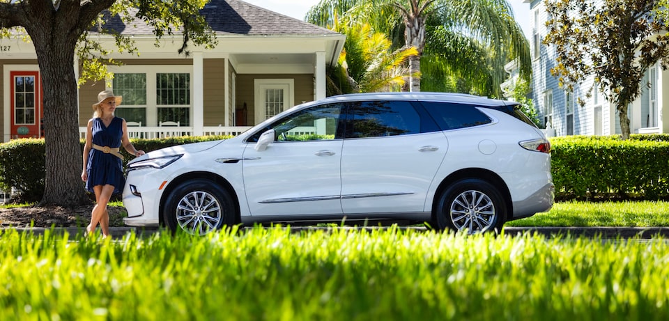 Side view of a white SUV surrounded by grassy plain with a woman in front of It