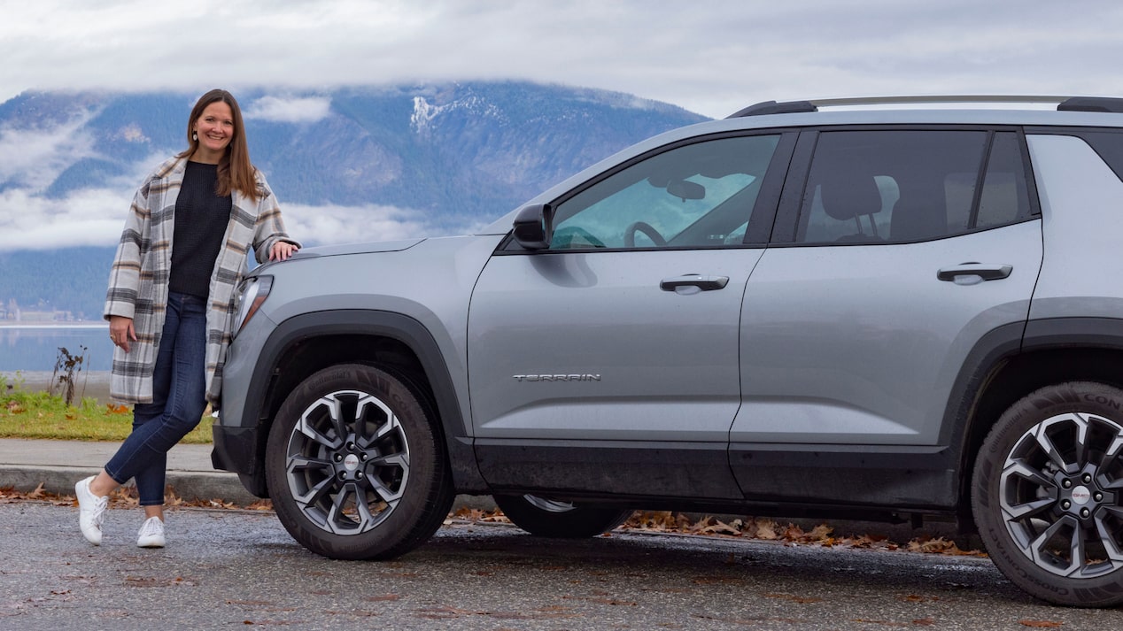 Sarah H. stands smiling next to her gray GMC Terrain, parked by a lake with mountains behind it.