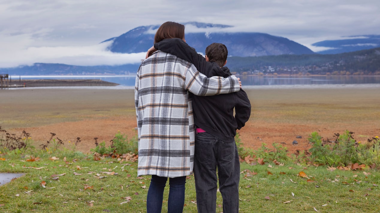 Sarah and her son in coats embrace, looking at a vast lake and distant mountains under a cloudy sky.