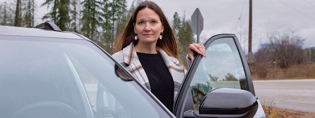 Sarah H. stands smiling by the open driver's door of her dark GMC Terrain with trees in the background.
