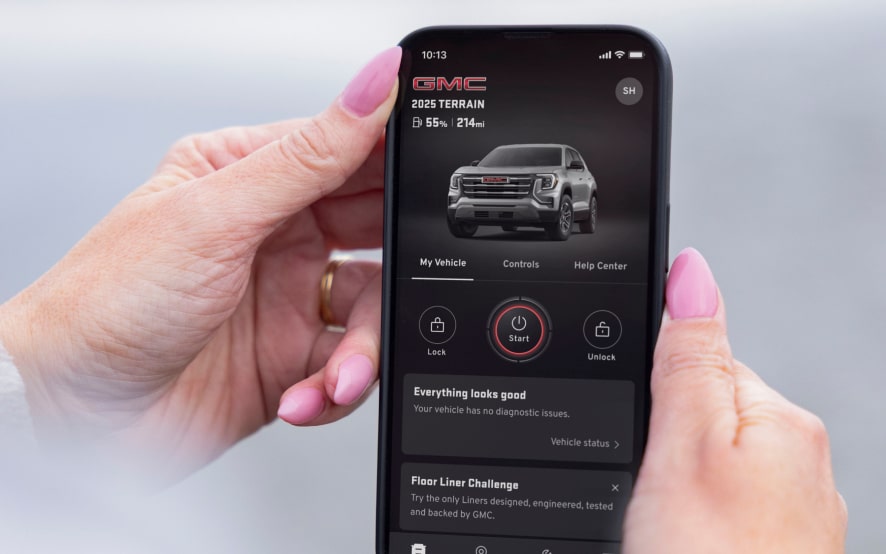 Hands hold a smartphone displaying the GMC app with a 2025 GMC Terrain model and vehicle controls.