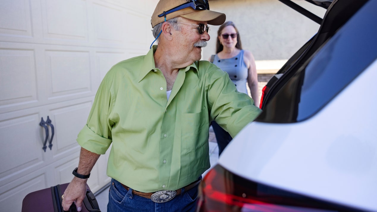 Andrew K. loads a suitcase into the open trunk of a white Cadillac XT6 while Sue K. stands nearby.