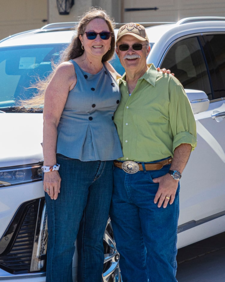 Andrew K. and Sue K. stand smiling in front of their white Cadillac XT6.