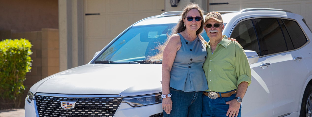 Andrew K. and Sue K. smile while standing in front of their white Cadillac XT6 with a garage in the background.