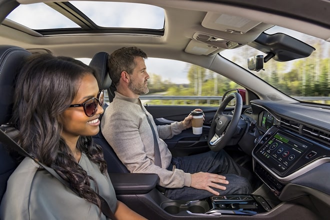 A man sitting in the driver's seat and holding a coffee with his left hand and a woman sitting in the passenger seat.