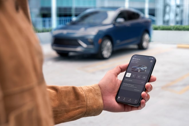 GET STARTED WITH V2H A hand holds a smartphone displaying the Buick mobile app with remote vehicle controls while a blue ENVISTA 2026 is parked in the background of an outdoor rooftop setting.